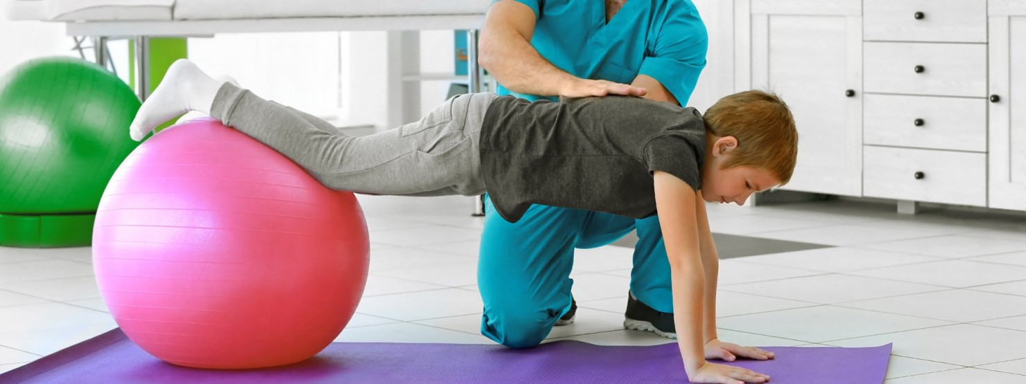 Ein Junge turnt mit einem rosa Gymnastikball und wird von einem Therapeuten unterstützt.