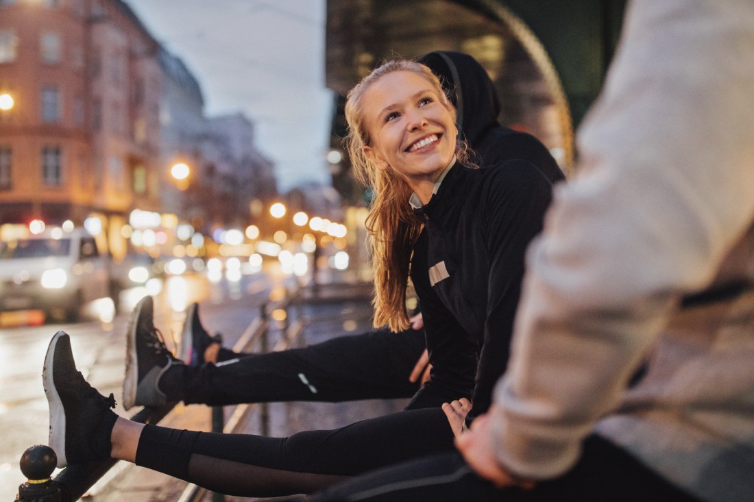 Eine junge Frau, Mitte 20, und zwei weiteren Personen stretchen ihre Beine an einem Geländer. Sie befinden sich zum Abend in urbanem Raum und sind guter Laune.
