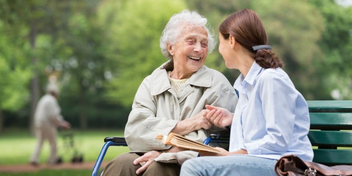Eine Seniorin sitzt mit einer etwa 40 jährigen Frau auf einer Bank. Beide reichen sich die Hände und lächeln sich an.