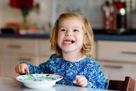 Ein etwa 3-jähriges Mädchen sitzt mit einem Teller am Tisch und streckt die Zunge raus.