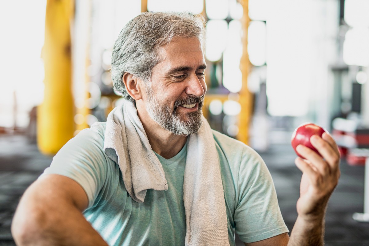 Etwa 55-jähriger Mann in einem Fitnessstudio betrachtet lächelnd einen Apfel in seiner Hand.
