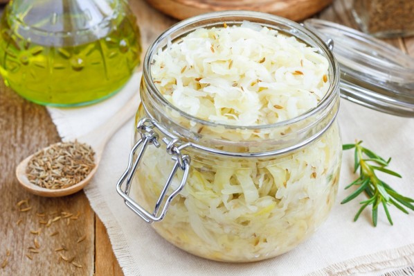 Auf einem Tisch steht ein offenes Glas selbstgemachter Sauerkraut. Daneben liegt ein Löffel mit Kümmel und ein Rosmarinzweig.