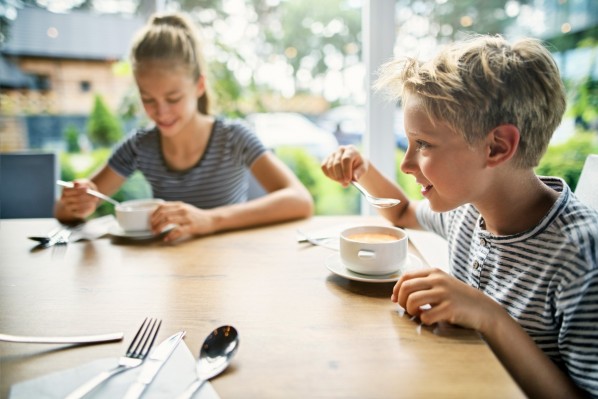 Zwei Teenager in gestreiften T-Shirts sitzen gut gelaunt am Küchentisch. Vor ihnen stehen Tassen voller Kartoffelsuppe, die sie mit einem Löffel essen.