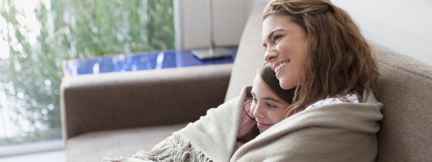Eine Mutter und ihre circa 6-jährige Tochter sitzen gemeinsam auf der Couch. Sie sind in eine Decke gewickelt.