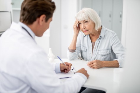 Eine Seniorin sitzt im Behandlungszimmer einer Arztpraxis. Ihr gegenüber sitzt ein Arzt im Gespräch der mitschreibt. Die Seniorin fässt sich während des Gesprächs mit der rechten Hand an die Schläfe.