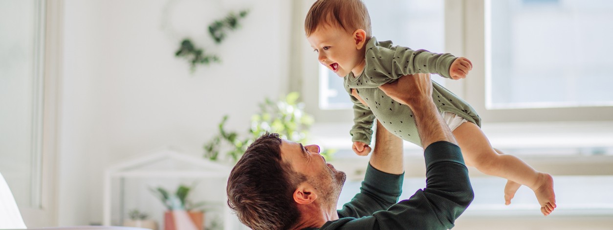 Ein Vater hält spaßig sein Baby mit Neugeborenenakne in die Luft.
