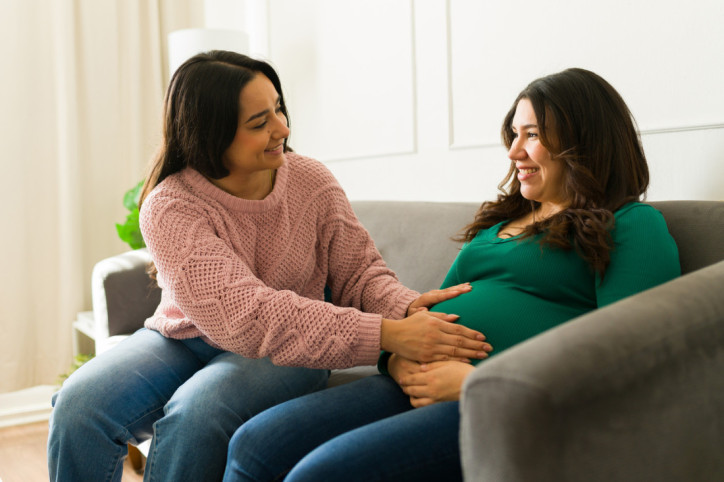 Schwangere Frau sitzt auf dem Sofa und spricht mit ihrer Doula, die zu einem  Hausbesuch vor Ort ist.