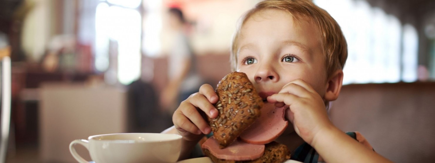 Ein etwa 3 jähriges Kind sitzt am Tisch und beißt von einem belegten Wurstbrötchen ab.