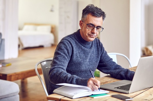 Ein Mann um die 50 sitzt am Schreibtisch vor einem Laptop und schreibt in ein Notizbuch.