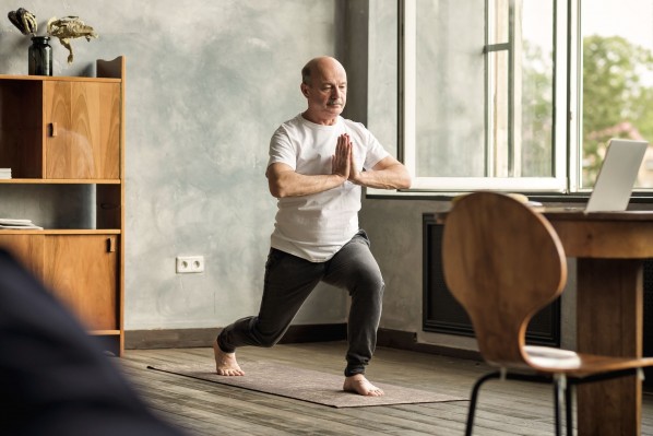 Senior bei einer Meditationsübung. Er steht dabei in seinem Wohnzimmer auf einer Yogamatte.