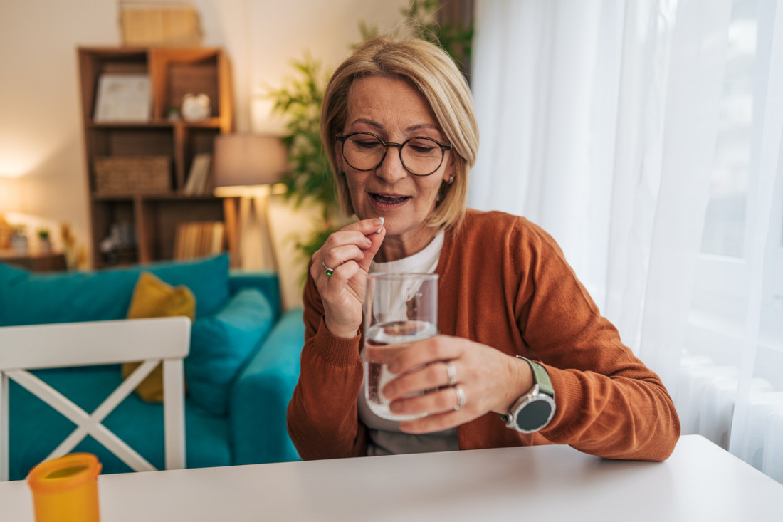 Senior woman sitting at the table in her living room, taking a pill with a glass of water, feeling unwell and seeking relief through medication in a bright, comfortable space