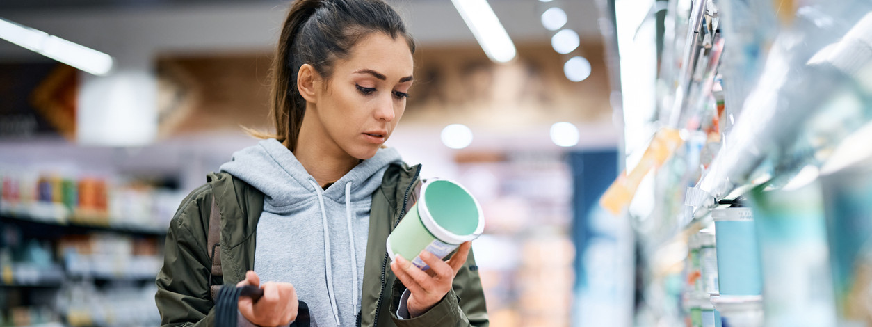 Eine etwa 25-jährige Frau steht im Supermarkt vor dem Produktregal und liest die Rückseite eines Produktes.