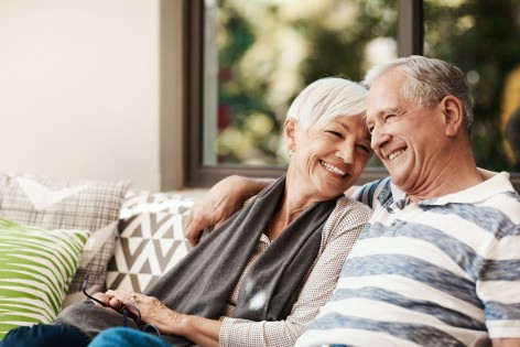 Ein Seniorenpaar sitzt Arm in Arm auf einem Sofa. Beide lächeln.