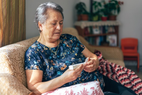 Eine etwa 80-jährige Frau sitz auf ihrem Sofa und betrachtet die Tablettendose in ihren Händen.