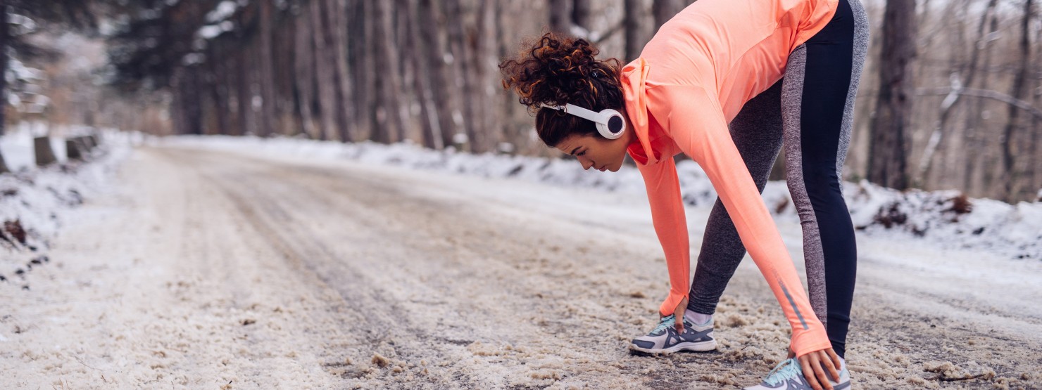 Eine etwas 30-jährige Frau steht an einer beschneiten Straße. Sie trägt Kopfhörer und dehnt ihre Beine.