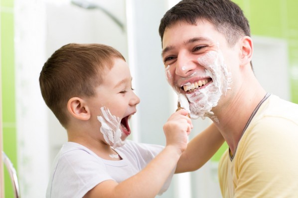 Ein Vater steht mit seinem etwa 5 jährigen Sohn im Bad. Der Junge rasiert seinen Vater im Gesicht. Beide haben Rasierschaum im Gesicht und lachen ausgiebig.
