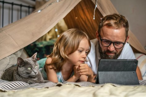 Ein Vater liegt mit seinem Kind auf einer Decke. Beide schauen gespannt auf ein Tablet.