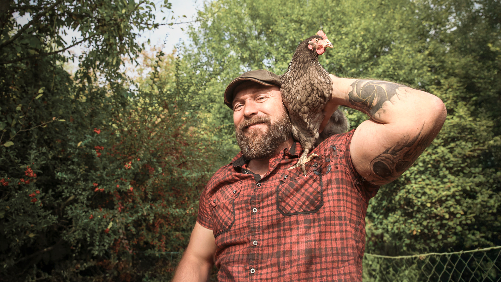 Ein kräftiger, tätowierter Mann, etwa Mitte 40, steht lächelnd im Garten und hat ein Huhn auf der Schulter. - AOK Sachsen-Anhalt begleitet mit starken Leistungen.