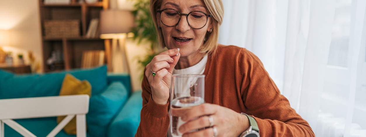 Senior woman sitting at the table in her living room, taking a pill with a glass of water, feeling unwell and seeking relief through medication in a bright, comfortable space