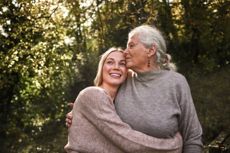 Eine etwa 25-jährige Frau und ihre etwa 75-jährige Großmutter stehen gemeinsam draußen im Grünen. Die junge Frau hat die Arme um die Mitte ihrer Großmutter geschlungen und lächelt glücklich. Ihr Blick schweift in die Ferne. Die Großmutter berührt sanft mit der Seite ihres Gesichts die Stirn der jungen Frau und hat ihre Hand sanft auf ihren Rücken gelegt.