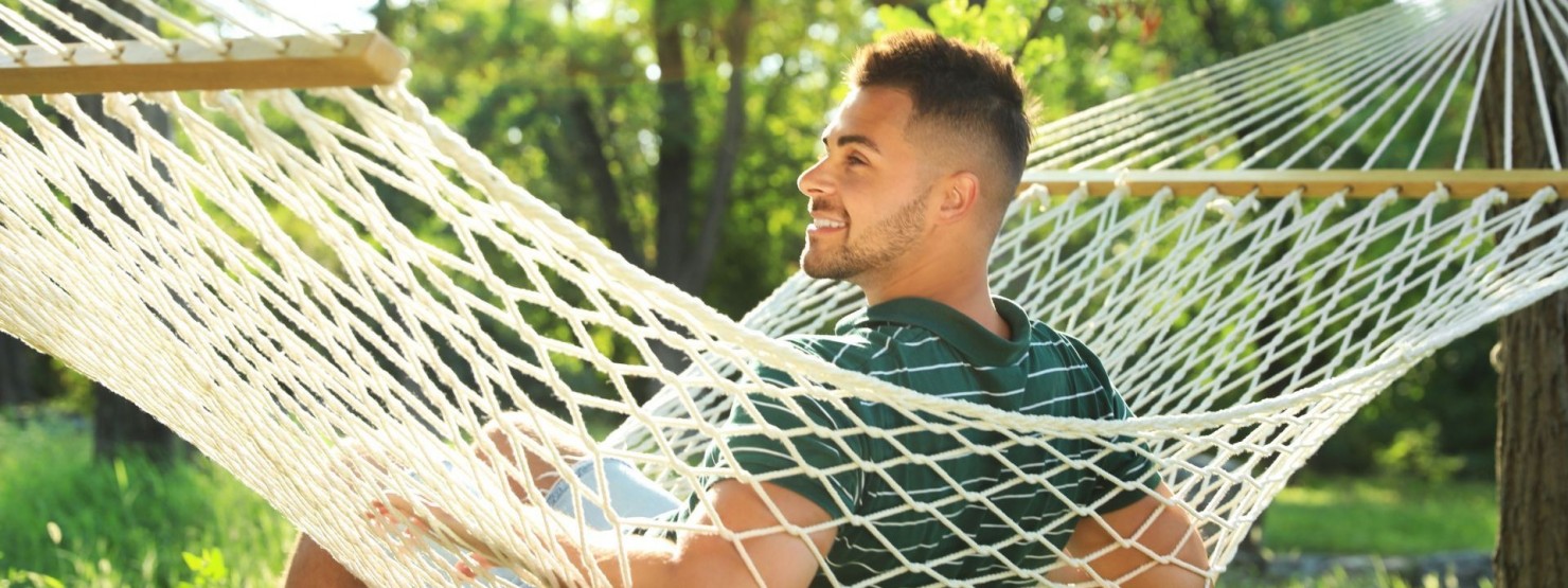 Ein etwa 30 jähriger Mann sitzt im Garten seitlich auf einer Hängematte. Die Sonne scheint und er lächelt.