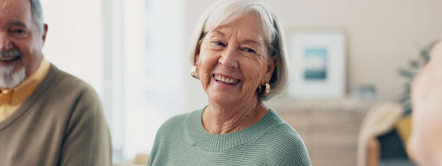 Zwei Seniorinnen sitzen auf einer Couch im Wohnzimmer und trinken gemeinsam Kaffee. Beide sind zueinander gedreht und lächeln sich an.
