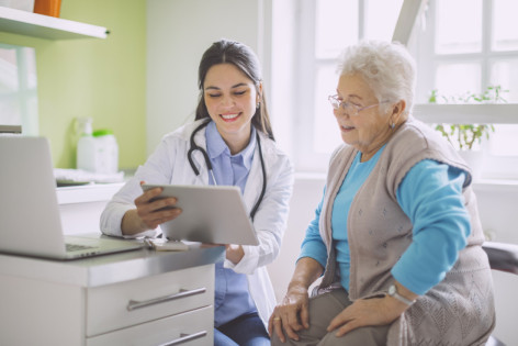 Eine Ärztin zeigt einer Seniorin verschiedene Toilettenhilfen auf einem Tablet.