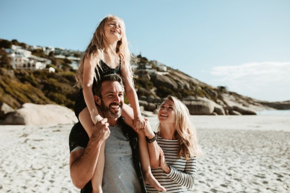 Family of three on a beach vacation. Father giving daughter ride on shoulders as they walk at the seashore with mother.