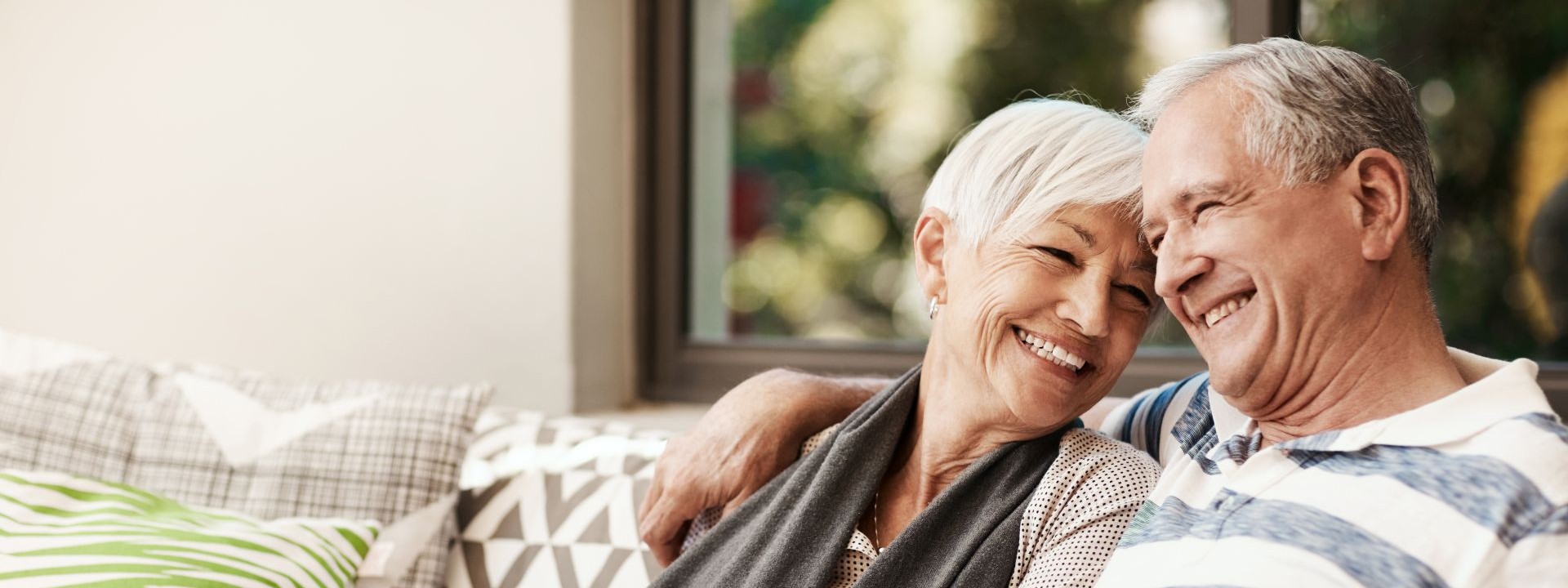 Ein Seniorenpaar sitzt Arm in Arm auf einem Sofa. Beide lächeln.