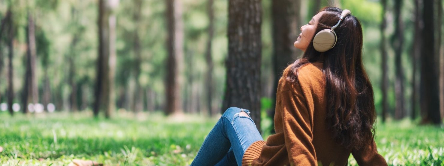 Eine etwa 25 jährige Frau sitzt mit Kopfhörern im Wald und genießt die Sonnenstrahlen.