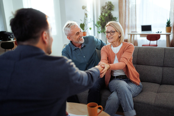 In a cozy living room, a mature couple shakes hands with a male insurance agent, finalizing an agreement. Smiles and a sense of satisfaction reflect a successful consultation and mutual trust.