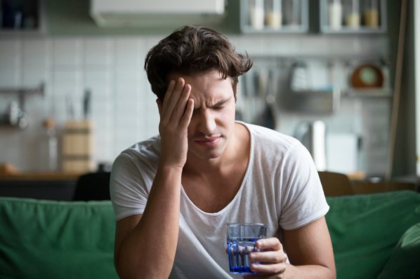 Ein junger Mann hat Kopfschmerzen und trinkt ein Glas Wasser.