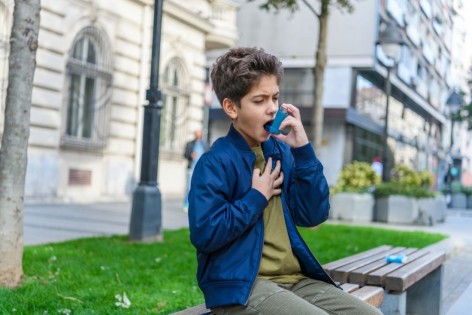 Ein ca. 8-jähriger Junge sitzt draußen auf einer Bank und benutzt sein Asthma-Spray