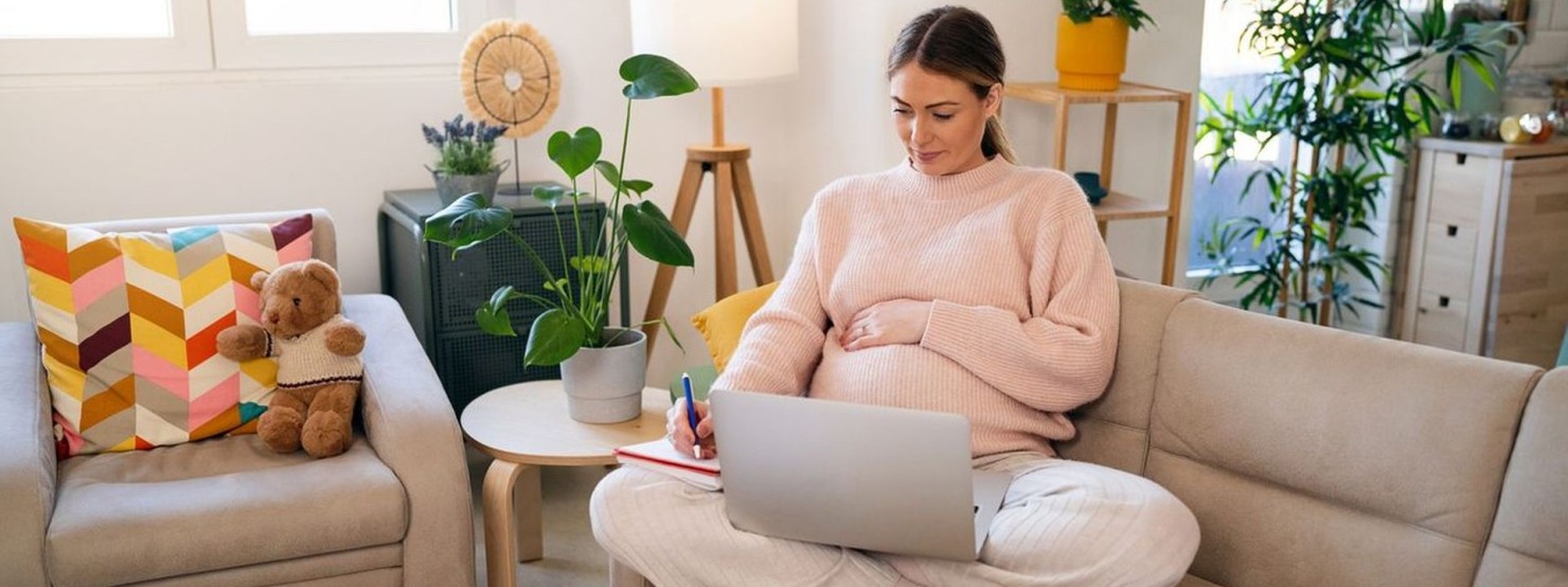 Eine circa 25-jährige, schwangere Frau sitzt mit Laptop auf der Couch.