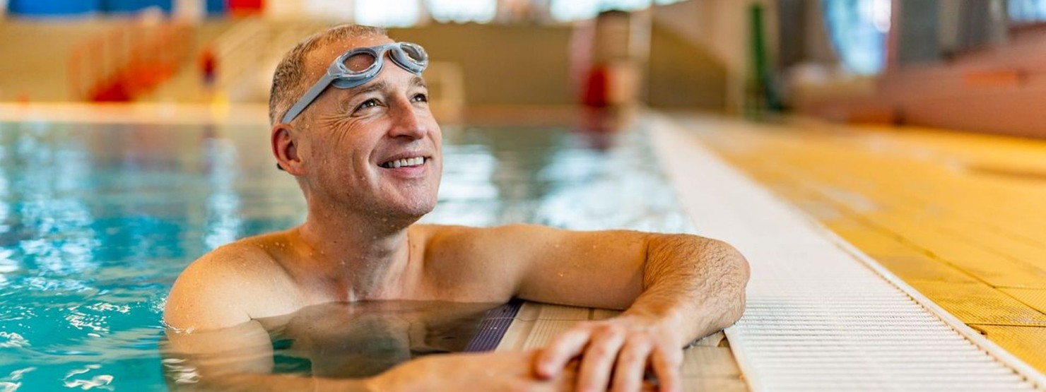 Ein etwa 55-jähriger Mann wartet am Beckenrand eines Schwimmbads und lächelt.
