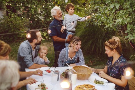 Mehrere Generationen einer Familie befinden sich zu einem sommerlichen Essen draußen in einem Garten. Der Großvater, etwa 65 Jahre, hat seinen Enkelsohn, etwa 4 Jahre, auf dem Arm und inspiziert mit ihm die zwischen den Bäumen gespannte Lichterkette. Zwei Frauen und ein Mann im Alter von etwa 30 bis 40 sitzen am Tisch in entspannter Atmosphäre beim Essen und Unterhalten. Der Mann hat ein weiteres, etwa zweijähriges Kleinkind auf dem Schoß und blickt zu Großvater und Kind hinüber.