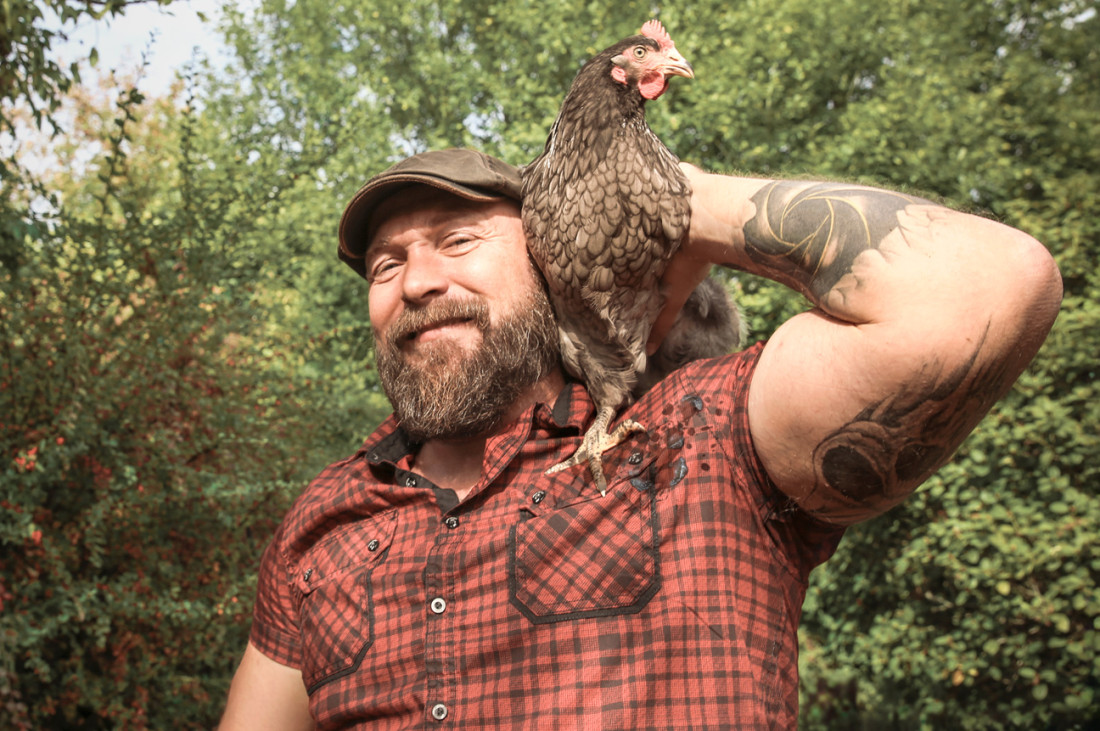 Ein kräftiger, tätowierter Mann, etwa Mitte 40, steht lächelnd im Garten und hat ein Huhn auf der Schulter. - AOK Sachsen-Anhalt begleitet mit starken Leistungen.