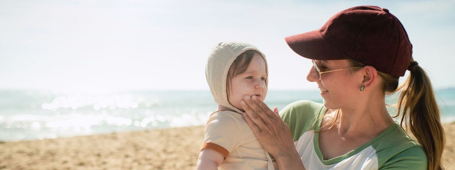 Eine junge Frau trägt ihr Kleinkind auf dem Arm und cremt es im Gesicht ein. Beide befinden sich am Strand und Meer.