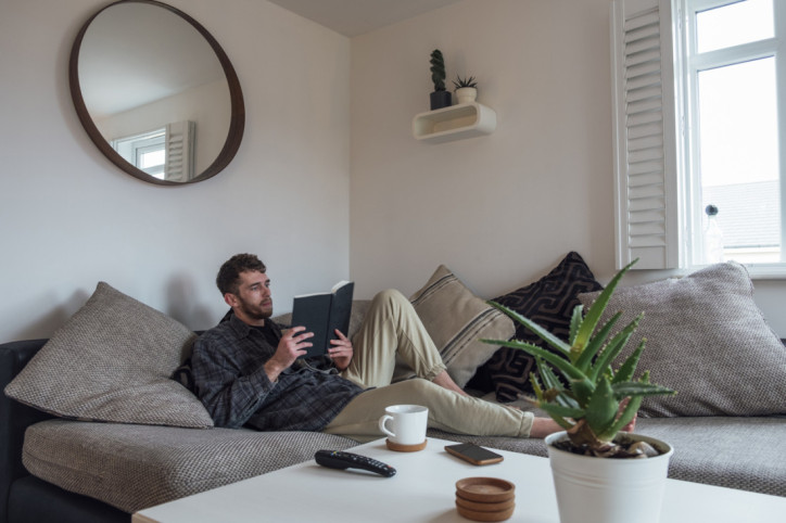 A caucasian man wearing casual clothing, sitting on a sofa in his apartment and reading a book.