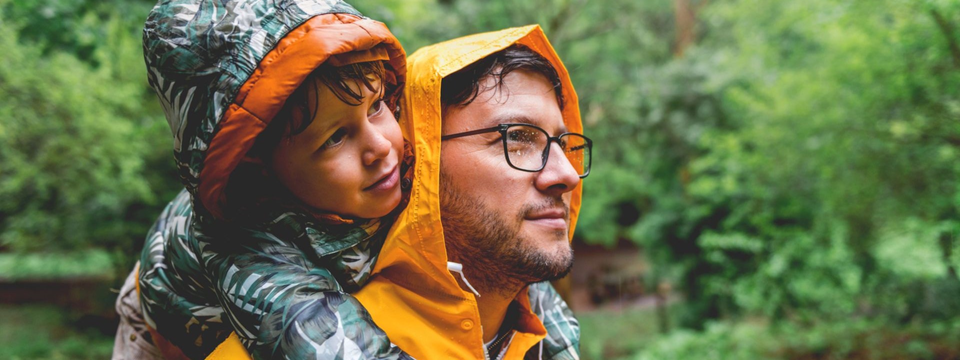 Vater ist mit seinem etwa 5-jährigen Sohn auf einem Waldspaziergang. Beide tragen aufgrund der Witterung einen Regenmantel.