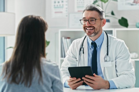 Eine Patientin wird von einem Arzt beraten. Dabei hält er ein Tablet in der Hand und lächelt die Patientin an.