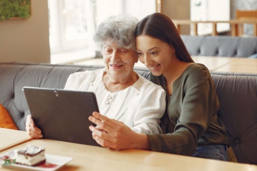 Eine etwa 39 jährige Frau schaut gemeinsam mit einer Seniorin auf ein Tablet. Beide lächeln-