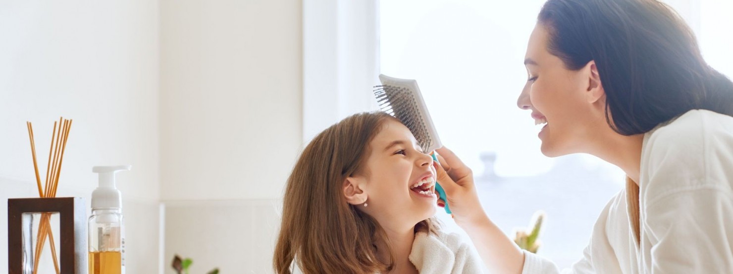 Eine Mutter sitzt mit ihrer Tochter im Ba. Beide tragen Bademäntel und lachen ausgiebig. Die Mutter kämmt ihrer Tochter die Haare.