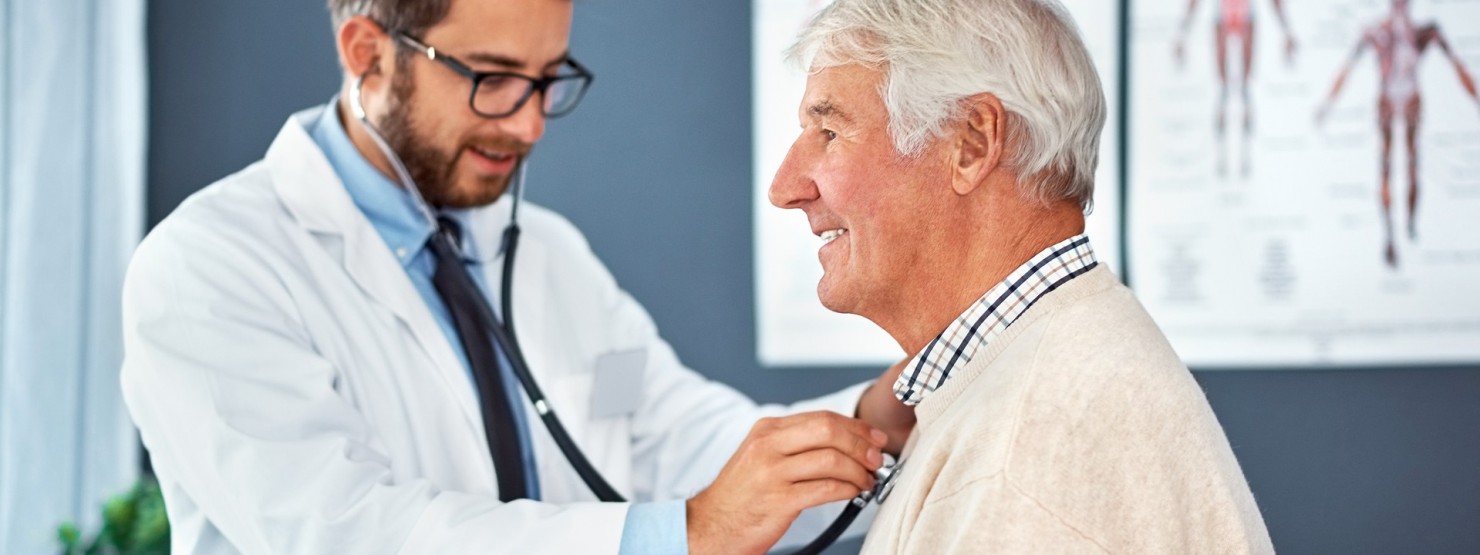 Ein etwas 35 jähriger Mann untersucht mit einem Stethoskop einen Senioren. Beide befinden sich in einem Behandlungsraum und lächeln.