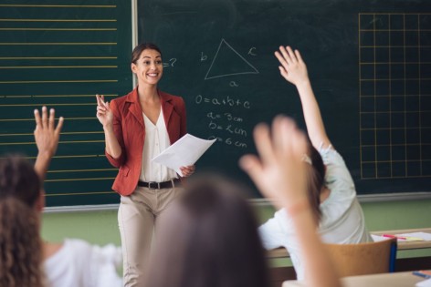 Eine Lehrerin steht vor der Tafel und spricht zur Klasse. Die Schülerkinder melden sich.