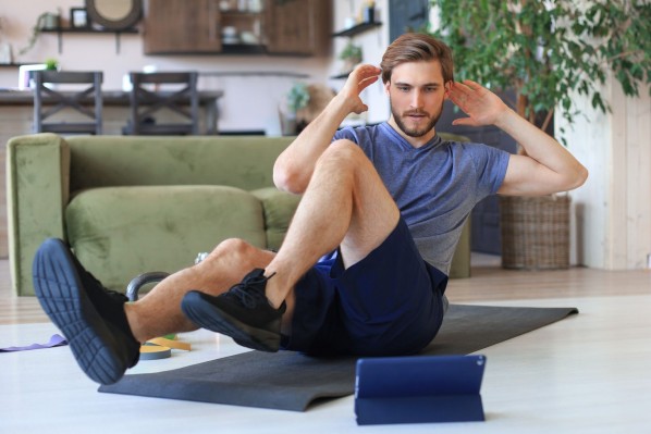 Ein etwa 25-Jähriger macht im Wohnzimmer auf einer Sportmatte Übungen eines online Kurses. 