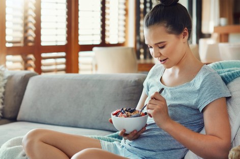 Junge Frau Anfang 20 sitzt auf dem Sofa und isst eine Schale mit Müsli