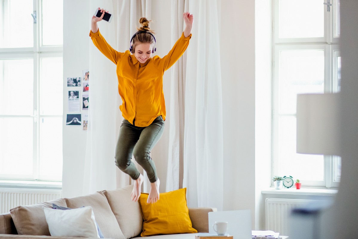 Eine etwa 23-Jährige springt freudig mit Kopfhörern auf ihrem Sofa herum.