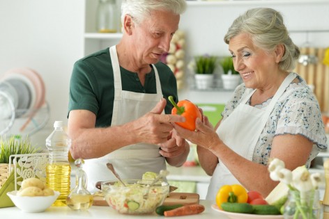 Ein Seniorenpaar steht gemeinsam in der Küche und kocht. Die Frau hält eine Paprika in der Hand. Beide lächeln.