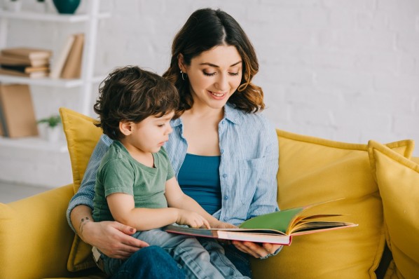 Eine Mutter hat ihr etwa zwei jähriges Kind auf dem Schoß. Beide sitzen auf dem Sofa und schauen sich ein Buch an.